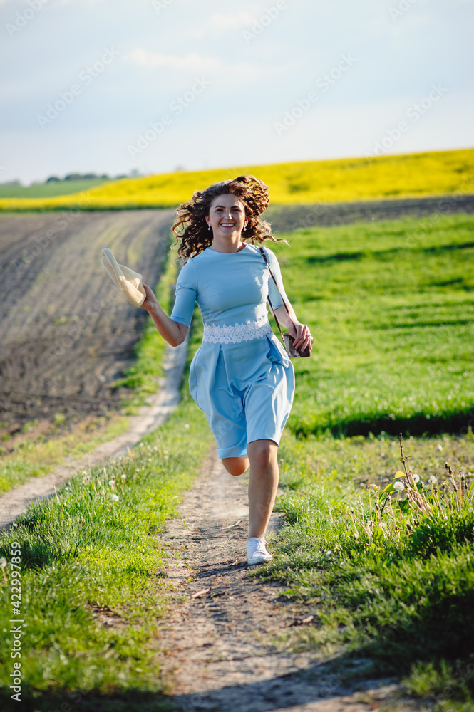 Happy curly brunette running across the field holding a straw hat. A young woman in a blue dress walks in nature. Green spring meadow. Happy curly brunette running across the field holding a straw hat. A young woman in a blue dress walks in nature. Green spring meadow.