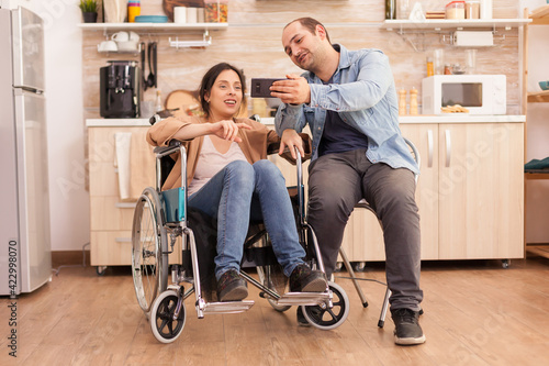 Cheerful disabled woman in wheelchair taking a selfie with husband in kitchen. Disabled paralyzed handicapped woman with walking disability integrating after an accident.