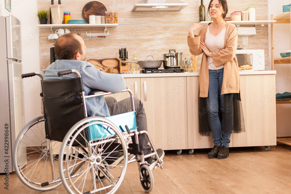 Unhappy man in wheelchair having a disagreement with wife in kitchen. Woman shouting at handicapped man. Disabled paralyzed handicapped man with walking disability integrating after an accident. Unhappy man in wheelchair having a disagreement with wife in kitchen. Woman shouting at handicapped man. Disabled paralyzed handicapped man with walking disability integrating after an accident.