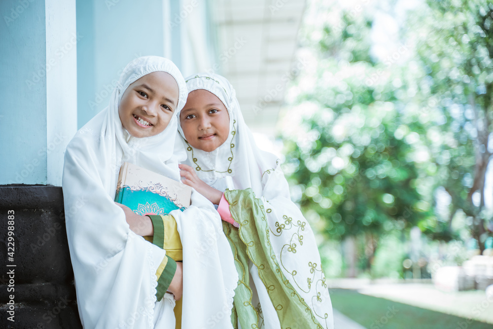 Portrait two Asian Muslim girls wearing mukena holding al quran book after salat together in the mosque