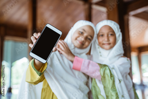 two Asian Muslim girls wearing mukena while hugging and showing telephone blank in the mosque photo