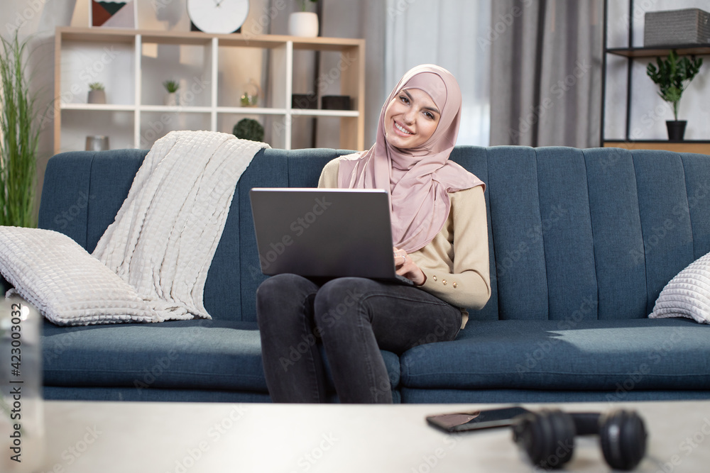 Distance work and freelance concept. Smiling beauty Muslim woman in hijab working remotely on laptop at home, sitting on sofa in living room. Online work and education. Distance work and freelance concept. Smiling beauty Muslim woman in hijab working remotely on laptop at home, sitting on sofa in living room. Online work and education.