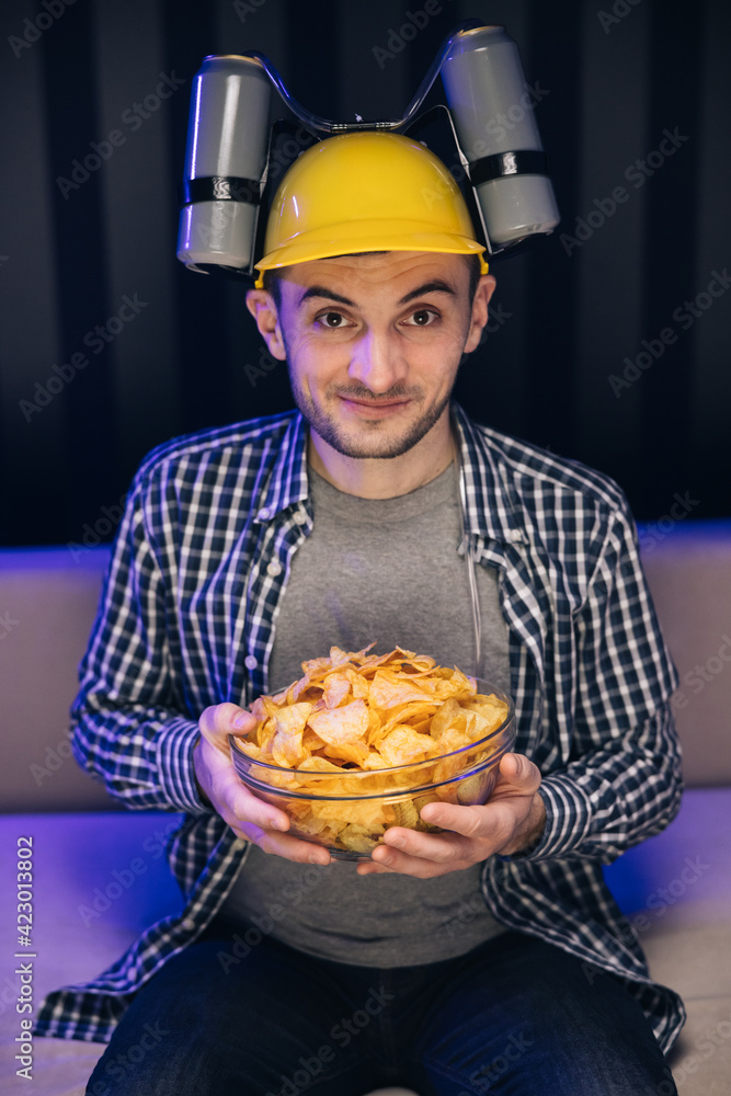 Handsome man with beer helmet on the head watching TV and eating potato chips on the couch at home. Young male sits on a sofa and eats snacks while watching a movie Handsome man with beer helmet on the head watching TV and eating potato chips on the couch at home. Young male sits on a sofa and eats snacks while watching a movie