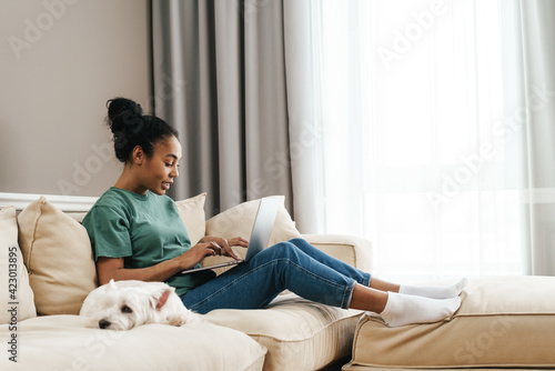 Smiling black woman using laptop while sitting with her dog on sofa