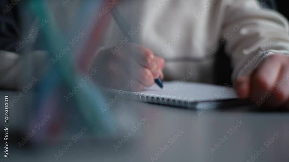 Close-up hands of unrecognizable pupil boy writing notes in paper workbook sitting at desk in classroom. Child schoolboy doing homework at home at table during daytime. Tracking shot in slow motion. Close-up hands of unrecognizable pupil boy writing notes in paper workbook sitting at desk in classroom. Child schoolboy doing homework at home at table during daytime. Tracking shot in slow motion.