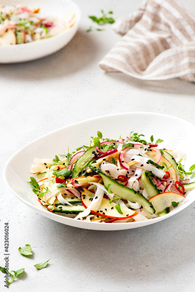 Rice noodle salad with cucumber, apple, onion, sesame seeds, and herbs in a white bowl on a light concrete background. Vegan salad Asian style. Vegetarian food. Vegetable salad with rice noodles Rice noodle salad with cucumber, apple, onion, sesame seeds, and herbs in a white bowl on a light concrete background. Vegan salad Asian style. Vegetarian food. Vegetable salad with rice noodles