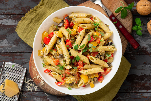 Pasta salad with baked vegetables. Penne pasta with baked peppers, eggplant, pesto and cheese in a white plate on a dark wooden table top view. Italian food. Rustic style.