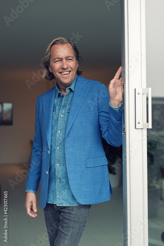 Smiling middle aged blonde man in blue jacket and white shirt standing in opened sliding door of living room.