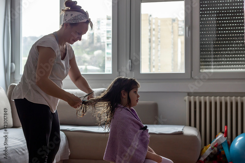 Young mother cutting her daughters hair at home during lockdown. Photo of cute girl with long blond hair having haircut at home. Mum doing haircut to her daughter at home during coronavirus outbreak.