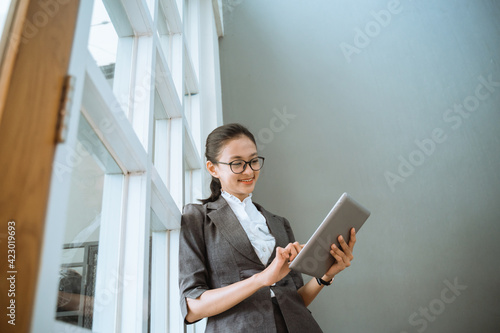 Portrait of young beautiful businesswoman looking at tablet standing and lean back in window office photo