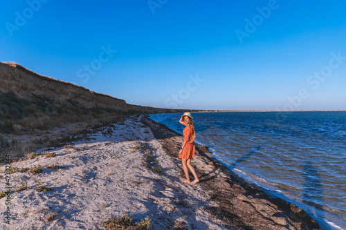 A beautiful girl walks along the seashore in a hat and in a red dress. Woman walking near the water on a warm day