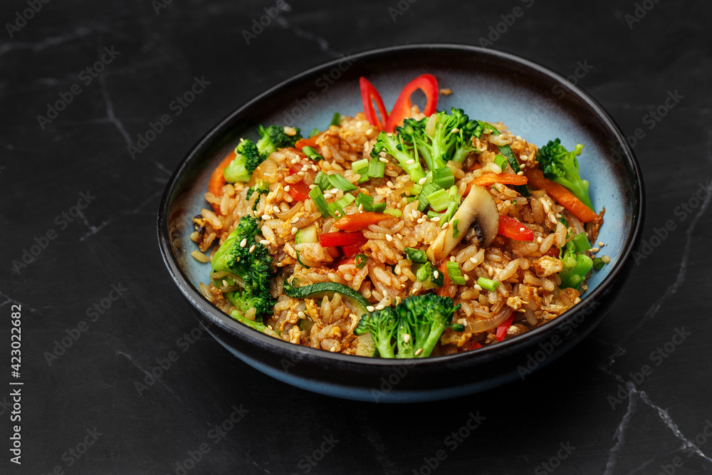 Asian rice with vegetables: zucchini, broccoli, red bel pepper, mushrooms, carrot, spring onion and sesame seeds. Dish isolated in a blue bowl, close-up on a black marble background. Asian cuisine. Asian rice with vegetables: zucchini, broccoli, red bel pepper, mushrooms, carrot, spring onion and sesame seeds. Dish isolated in a blue bowl, close-up on a black marble background. Asian cuisine.