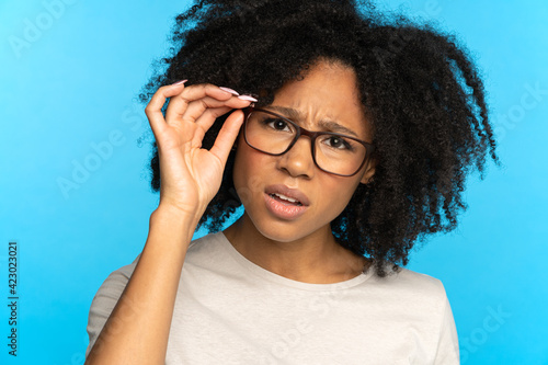 Bild auf Leinwand Studio portrait of unsure doubtful African American girl touching glasses, suspicious looking at camera, isolated on blue blank background