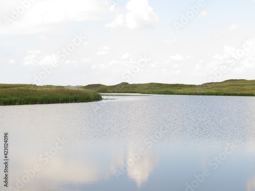 Small ripples on the water surface of the small lake.Cloudy warm summer day with a little breeze. Reflection of clouds in the water. Grass on the shore