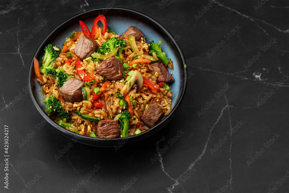 Asian rice with teriyaki beef and vegetables: zucchini, broccoli, pepper, mushrooms, carrot, onion and sesame seeds. Dish isolated in a blue bowl, close-up on a black marble background. Asian cuisine. Asian rice with teriyaki beef and vegetables: zucchini, broccoli, pepper, mushrooms, carrot, onion and sesame seeds. Dish isolated in a blue bowl, close-up on a black marble background. Asian cuisine.