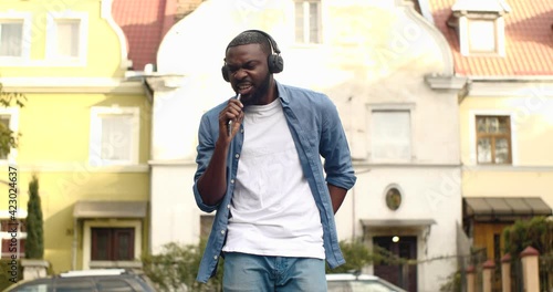 Cheerful Afro American Guy with Headphones Singing favourite Song in Smartphone. Having Positive Emotions Outdoors while Walking. Attractive Man listening to Music imagines himself on stage. Fun.