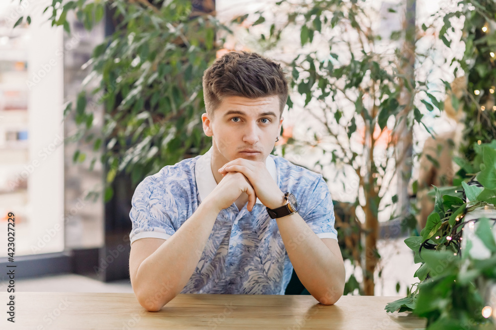 Fototapeta premium A handsome young man with an earring in his ear sits at the table with his hands folded and looks at the camera