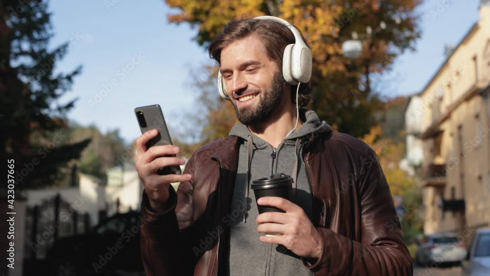 Handsome Smiling Bearded Guy with Headphones Typing on Mobile Phone Outdoors. Attractive Man Browsing on Social Networks while Resting in Sunny Autumn Day. Searching favourite Music on Smartphone. Handsome Smiling Bearded Guy with Headphones Typing on Mobile Phone Outdoors. Attractive Man Browsing on Social Networks while Resting in Sunny Autumn Day. Searching favourite Music on Smartphone.