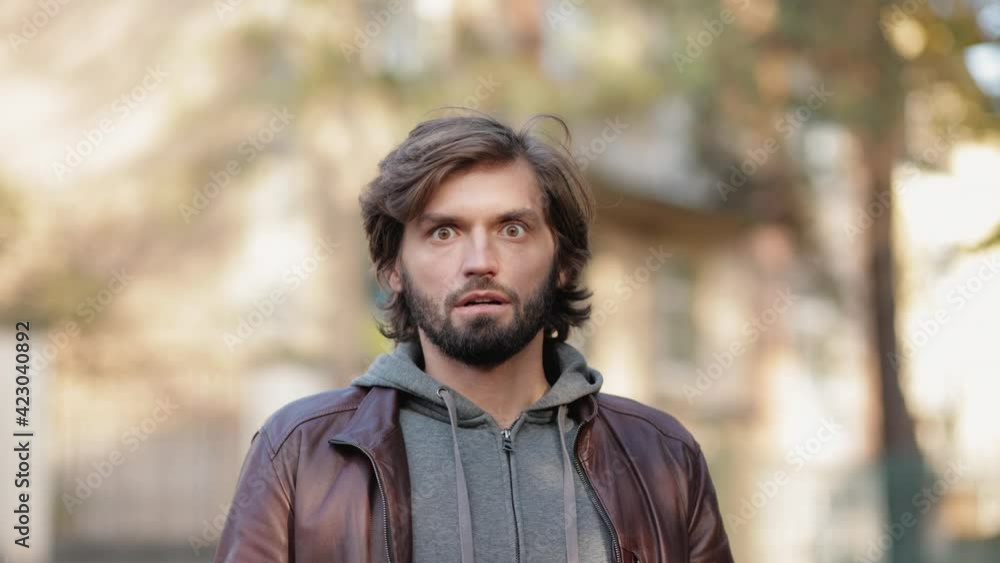 Portrait of Young Man Outdoors Shows Shocked Emotion looking at the Camera. Caucasian Bearded Man standing at the Park looking Intently wearing Casual Clothes. Surprised Face. Portrait of Young Man Outdoors Shows Shocked Emotion looking at the Camera. Caucasian Bearded Man standing at the Park looking Intently wearing Casual Clothes. Surprised Face.