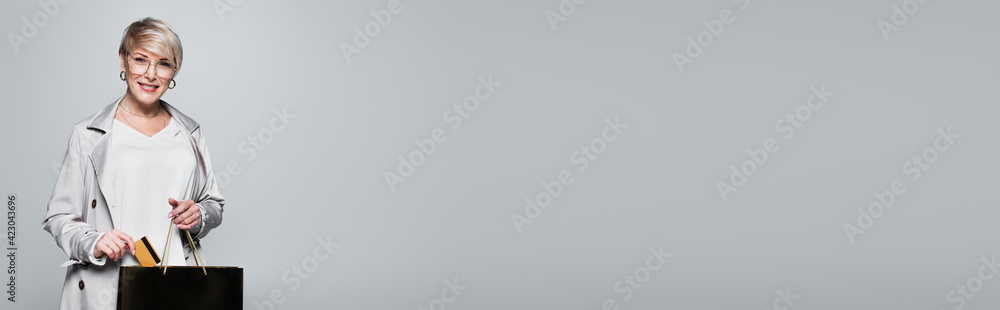 happy woman in stylish outfit putting credit card into shopping bag isolated on grey, banner happy woman in stylish outfit putting credit card into shopping bag isolated on grey, banner
