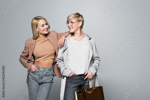trendy mother and daughter smiling at each other while posing with hands in pockets on grey