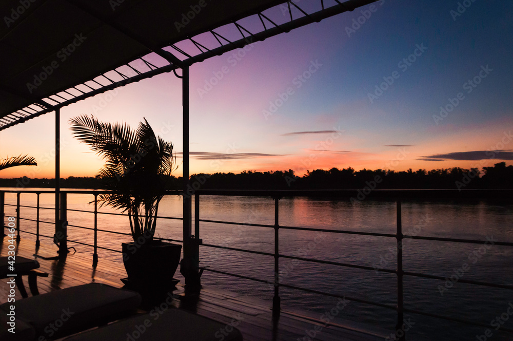 Abendstimmung auf dem Mekong. Blick von einem Flußschiff mit Reling, auf das Ufer mit der untergehenden Sonne