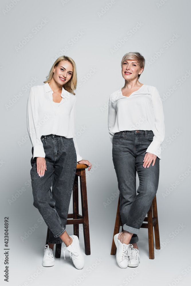 Obraz premium full length view of smiling mother and daughter in trendy clothes posing near high stools on grey
