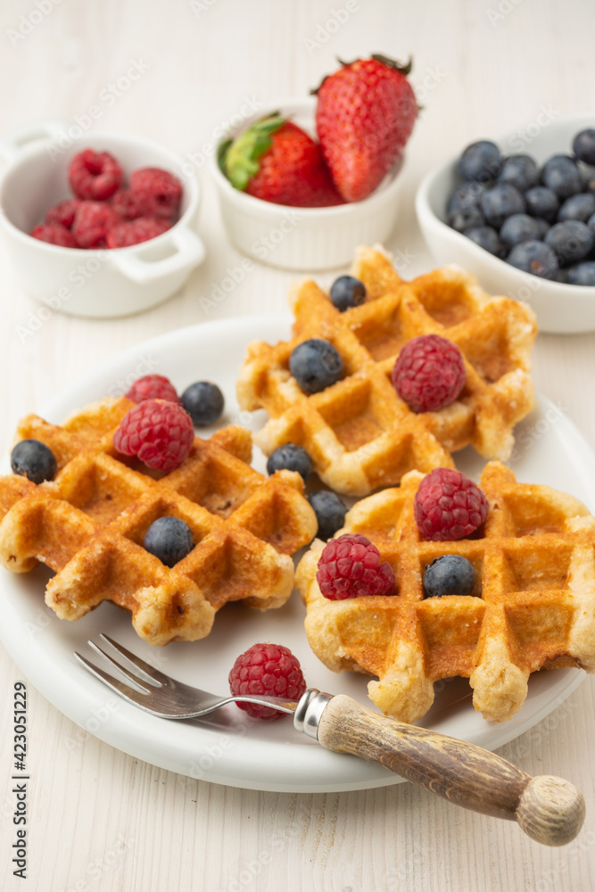 Top view of waffles with raspberries and blueberries, on plate with fork, and bowls with strawberries, raspberries and blueberries, selective focus, on white table, in vertical Top view of waffles with raspberries and blueberries, on plate with fork, and bowls with strawberries, raspberries and blueberries, selective focus, on white table, in vertical