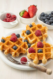 Top view of waffles with raspberries and blueberries, on plate with fork, and bowls with strawberries, raspberries and blueberries, selective focus, on white table, in vertical