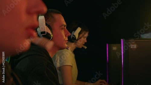 A team of two guys and girls play a cyber match. They are actively talking over microphones. Are in a dark room they are illuminated only by computer monitors