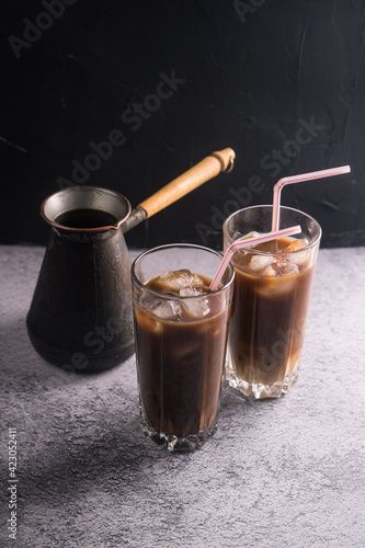 Two tall glasses of cold coffee with milk and ice with tubes on a dark background with a coffee maker. Copy space. photo