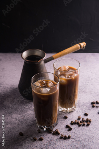 Coffee maker with two tall glasses of cold coffee with milk and ice on a dark background and scattered grains of natural coffee. Copy space. The concept of refreshing drinks for summer photo