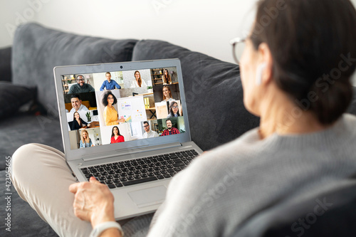 Contemporary senior woman lying on the comfortable sofa and using laptop for video connection with group of diverse people, back view an older female involved webinar, video conference