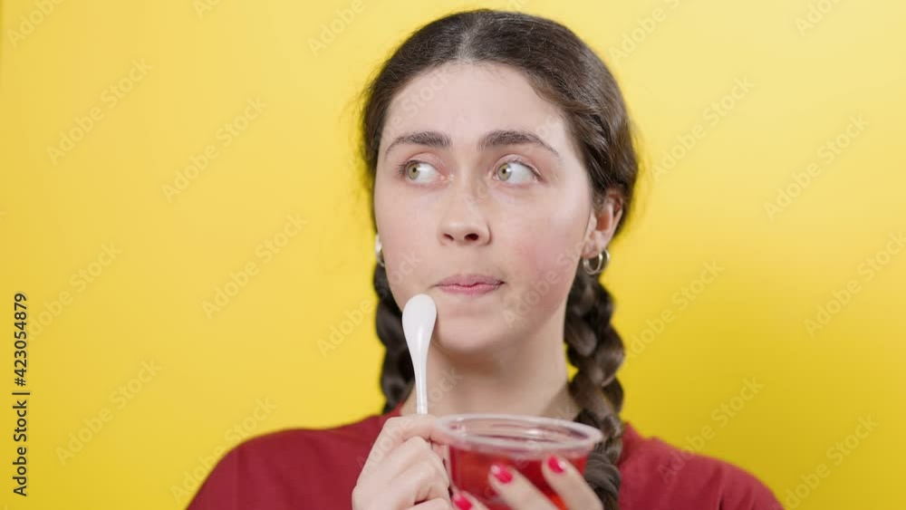 Portrait of a young smiling woman who sniffs a fragrant jar of jelly, and thoughtfully puts a spoon to her mouth. Yellow background. Diet Concept Portrait of a young smiling woman who sniffs a fragrant jar of jelly, and thoughtfully puts a spoon to her mouth. Yellow background. Diet Concept