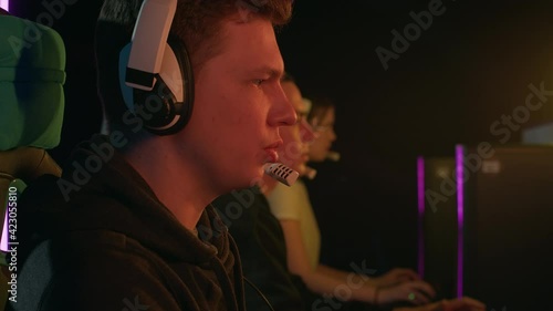 Medium shot of a man speaking into a microphone while playing a video game. It is illuminated by a monitor with overflowing colors. He plays with his team that sits next to. High quality 4k footage