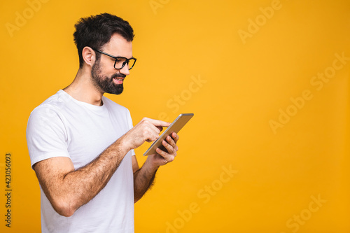 Happy young bearded man in casual standing and using tablet isolated over yellow background.