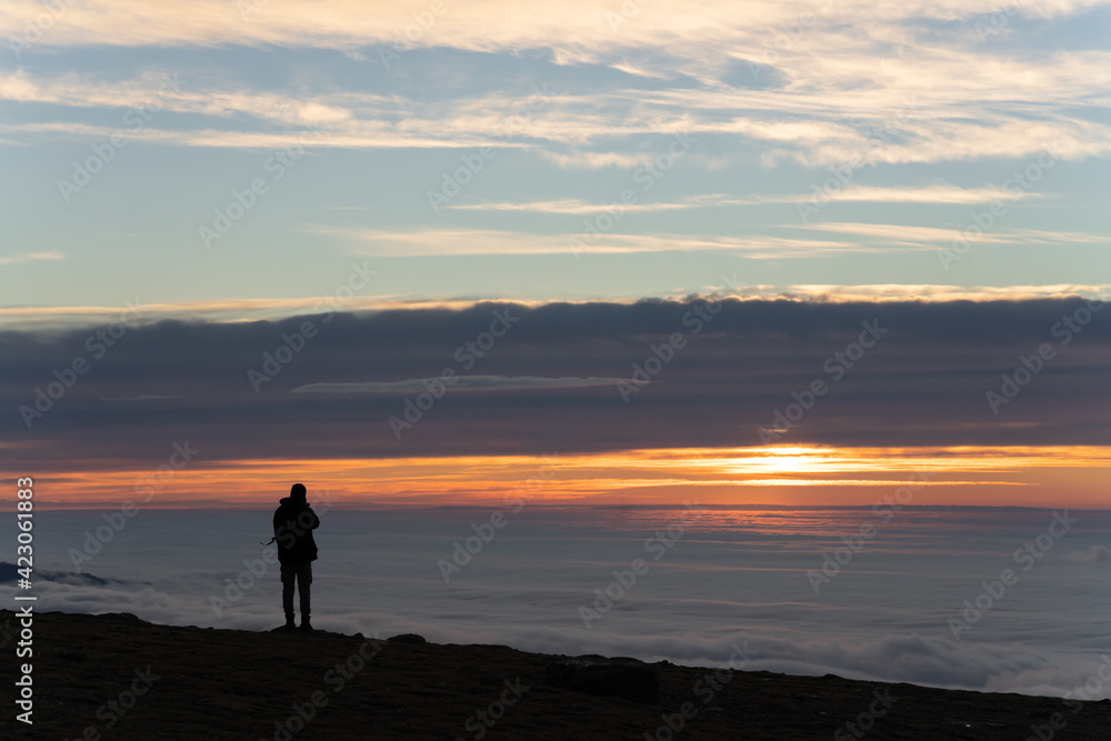 Back view of unrecognizable explorer male tourist in warm outerwear with backpack standing contemplating mountain under thick clouds at dawn Back view of unrecognizable explorer male tourist in warm outerwear with backpack standing contemplating mountain under thick clouds at dawn