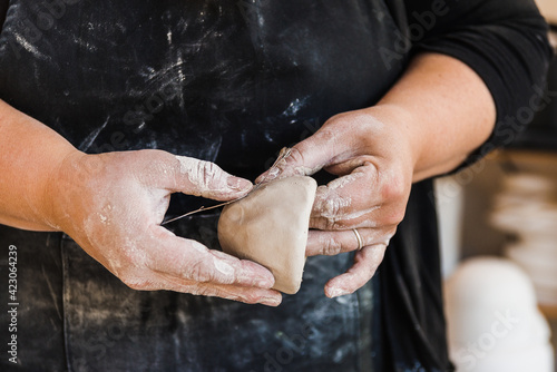 Crop anonymous master in dirty apron standing in workshop and shaping piece of clay marking it with flower in hands photo