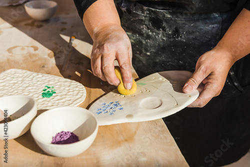 Crop anonymous female artisan in dirty black apron using sponge for filling in drawing with pigment on ceramic palette on table near bowls with paint in bright workshop photo