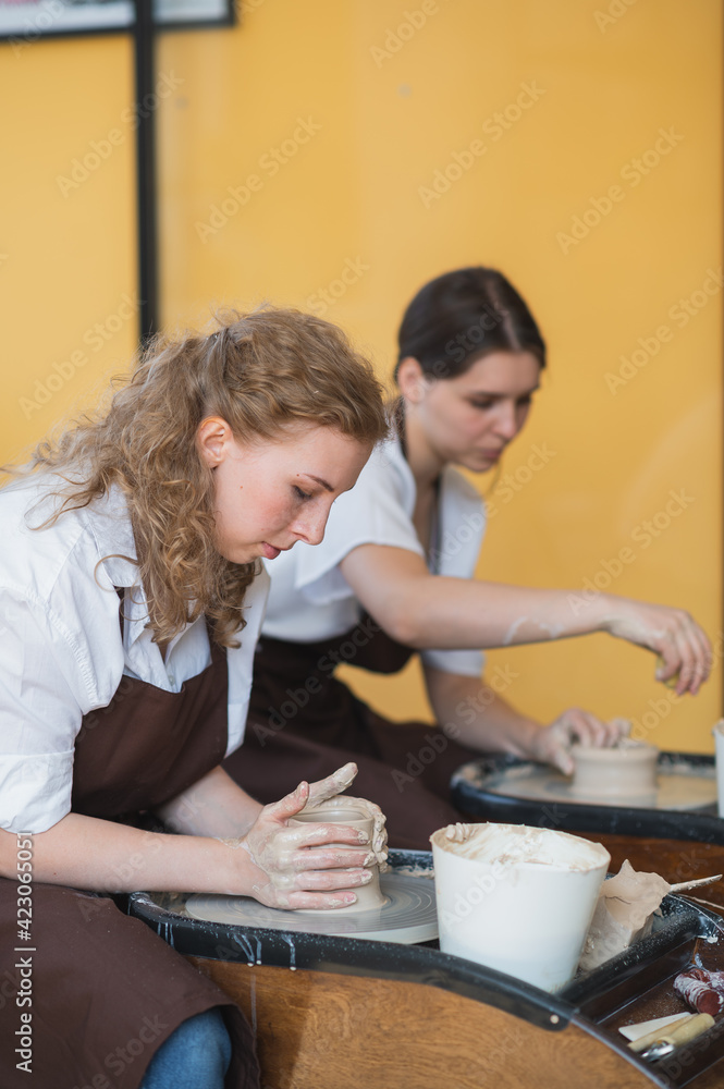 Young woman potter in apron at work. Craftsman artist shapes the jug with her hands and a special tool on pottery wheel. Young woman potter in apron at work. Craftsman artist shapes the jug with her hands and a special tool on pottery wheel.