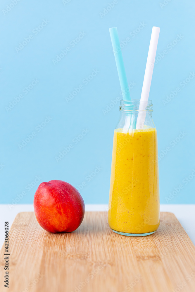 Fototapeta premium Smoothie or cocktail of nectarine, almond or oat milk and turmeric in a glass jar with two cocktail tubes. Juicy ripe nectarine on a cutting board on a blue background. Copy space, vertical.