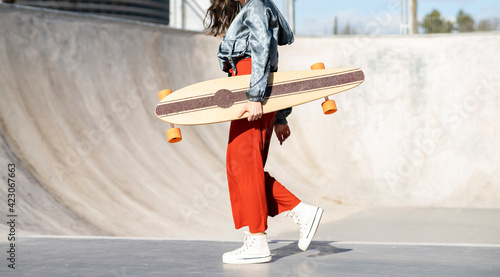 Side view of cropped unrecognizable happy young female in stylish apparel walking with longboard on concrete skate park