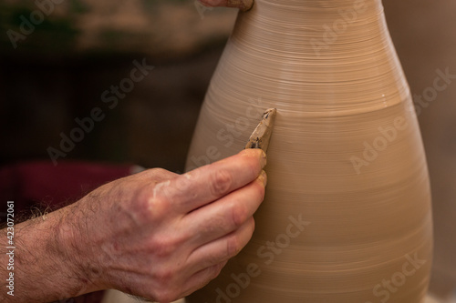Crop unrecognizable sculptor with equipment giving shape while sculpting with brown clay on throwing wheel