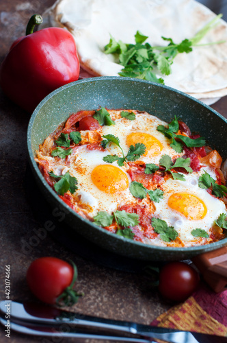 Traditional dish of israeli cuisine Shakshuka. Fried eggs with tomatoes, peppers, onions, cilantro and pita in a pan on dark brown background
