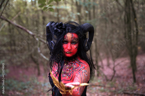Sexy Teufelin steht im Wald. Sie hat die Hörner auf dem Kopf, eine schwarze Corsage. sie ist mit der roten Farbe und der Muster angemalt.Sie hat lange, schwarze Haare.Überall ist der roter Rauch.