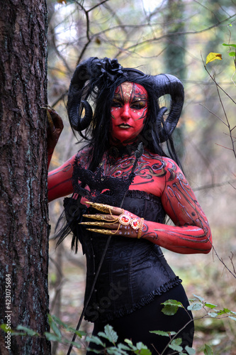 Sexy Teufelin steht im Wald. Sie hat die Hörner auf dem Kopf, eine schwarze Corsage. sie ist mit der roten Farbe und der Muster angemalt.Sie hat lange, schwarze Haare. Nah Portrait.