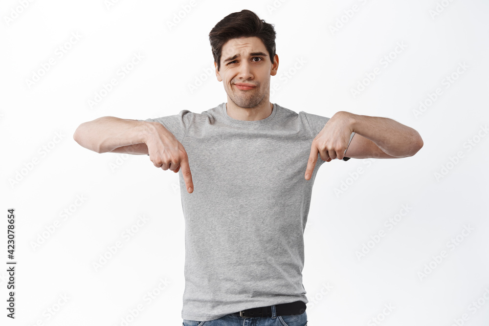 Image of skeptical brunette man smirk and frowns, pointing fingers down at something bad or average, have doubts, standing unamused against white background Image of skeptical brunette man smirk and frowns, pointing fingers down at something bad or average, have doubts, standing unamused against white background