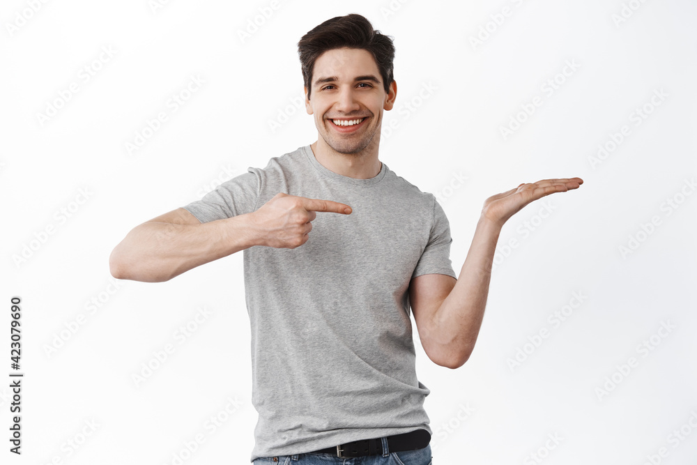 Image of handsome smiling man pointing at his empty hand, holding item on palm, display your product, demonstrate copy space, standing satisfied against white background Image of handsome smiling man pointing at his empty hand, holding item on palm, display your product, demonstrate copy space, standing satisfied against white background