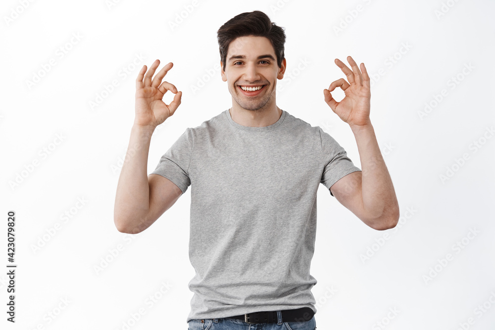 This is fine. Smiling handsome guy shows okay signs, reassure and give guarantees everything good, praise excellent job, standing over white background This is fine. Smiling handsome guy shows okay signs, reassure and give guarantees everything good, praise excellent job, standing over white background
