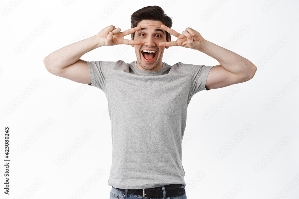 Excited happy man in t-shirt showing peace signs, v-sign kawaii over eyes and looking joyful, smiling, standing over white background Excited happy man in t-shirt showing peace signs, v-sign kawaii over eyes and looking joyful, smiling, standing over white background